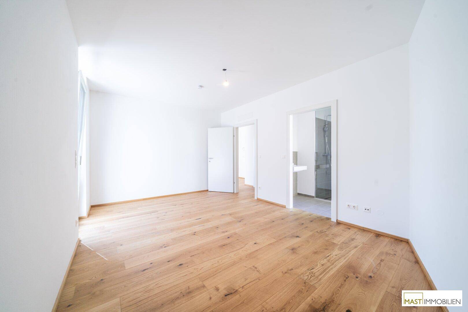 Heller Raum mit Holzboden und weiß gestrichenen Wänden, mit direktem Zugang zu einem Badezimmer.