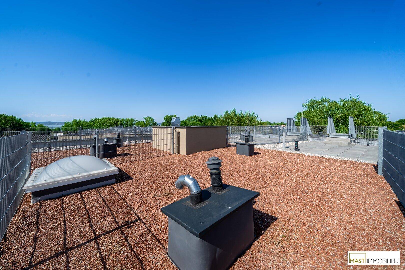 Teil der Dachterrasse mit Kiesbelag, Lüftungselementen und Blick auf die grüne Umgebung.