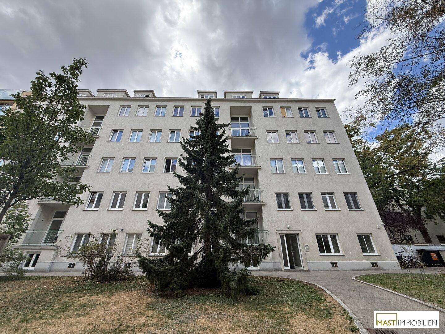 Mehrstöckiges Wohngebäude mit Balkonen und einem großen Nadelbaum im Vorgarten.