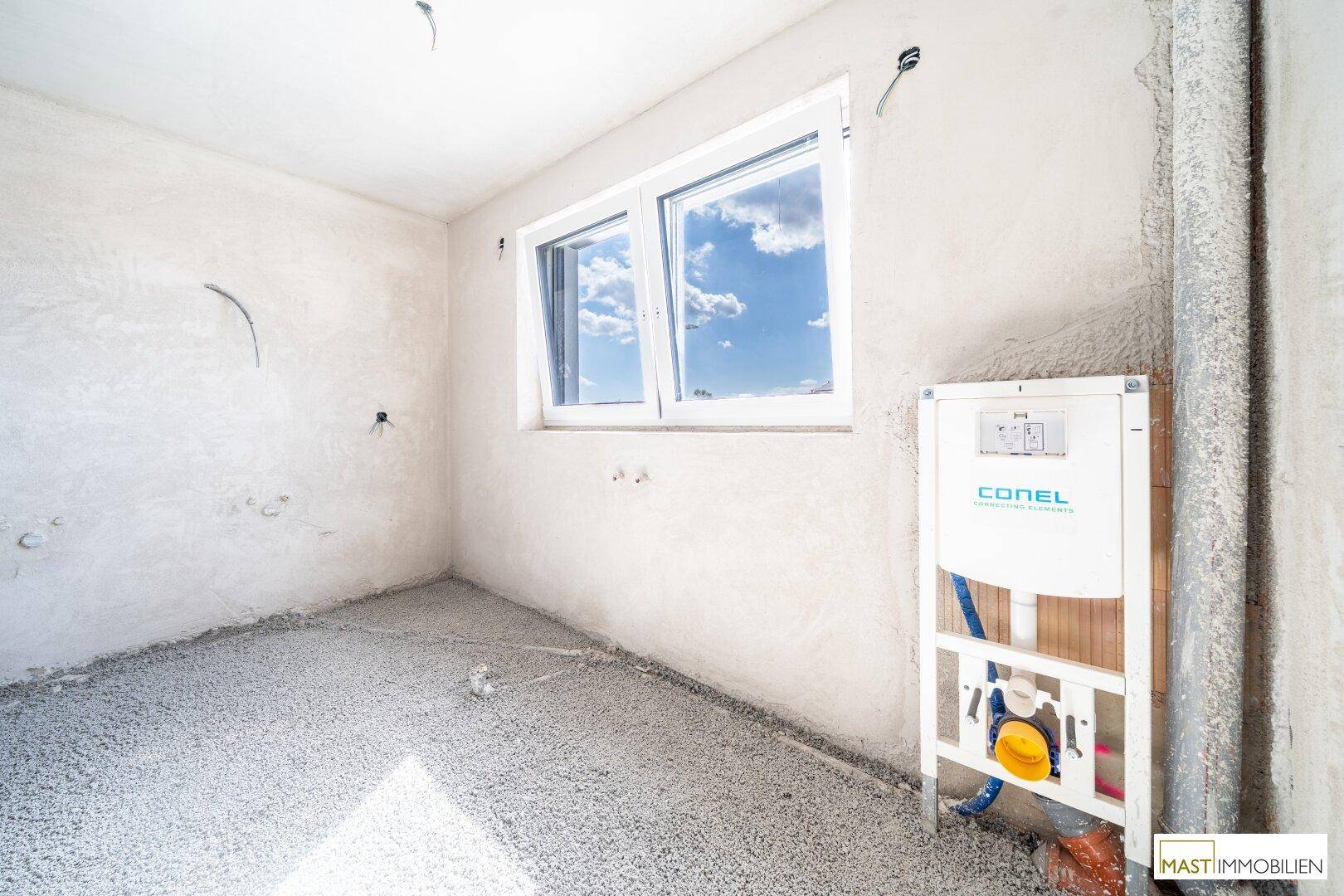 Unfertiges Badezimmer mit Fenster und vorbereiteter Installation für Toilette und Waschbecken.