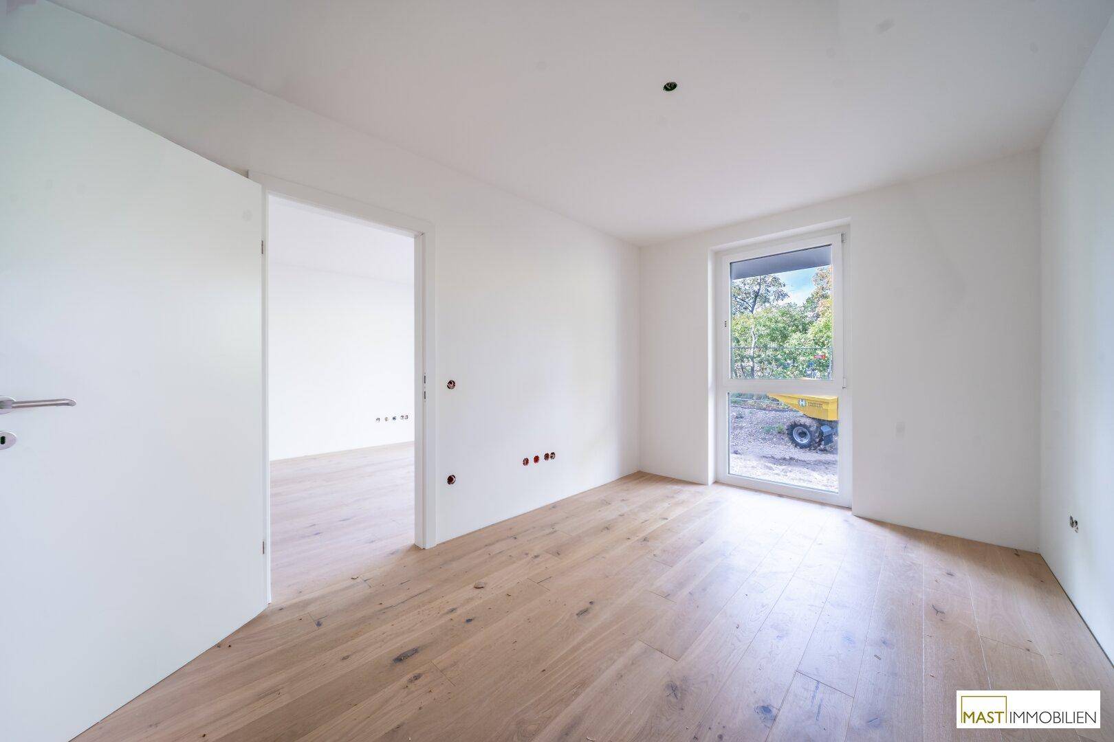 Helles Schlafzimmer mit Holzboden und Fensterblick auf die Baustelle und Bäume.