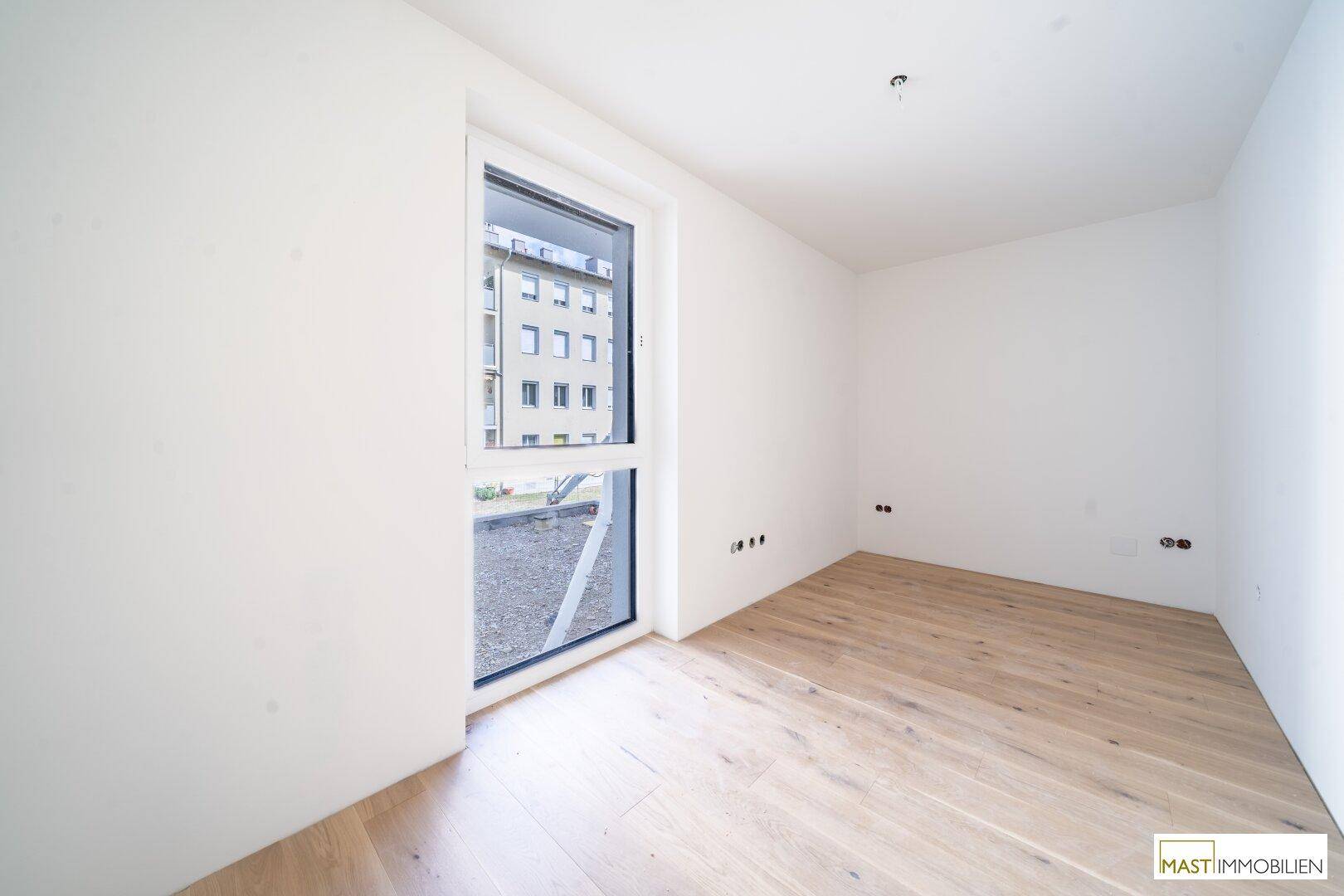 Helles Zimmer mit großem Fenster und Holzboden, bietet viel Tageslicht und eine neutrale Basis.