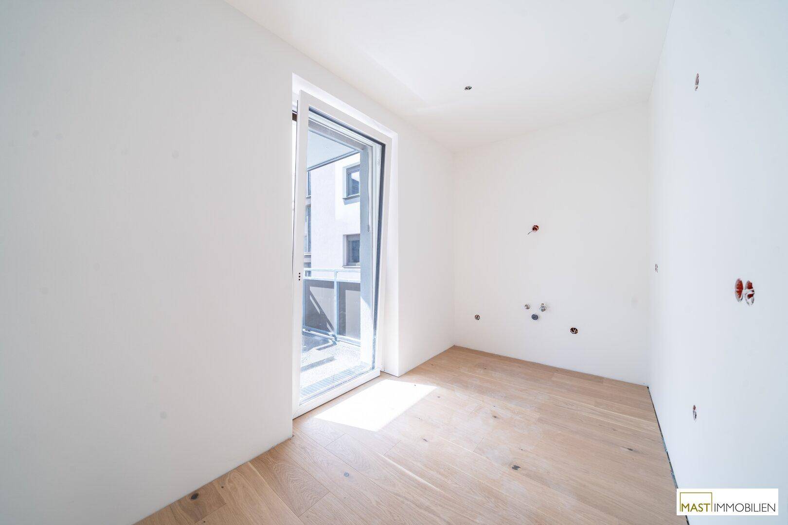 Helles Zimmer mit Holzboden und Zugang zu einem kleinen Balkon.