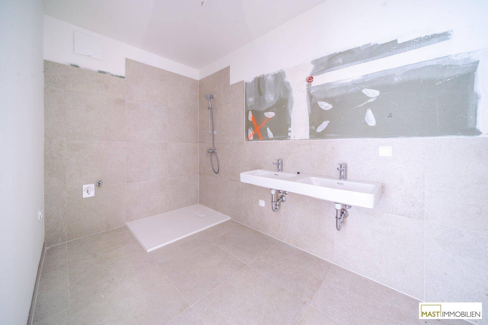 Modernes Badezimmer im Bau mit großen grauen Fliesen, einer begehbaren Dusche und einem Doppelwaschbecken.
