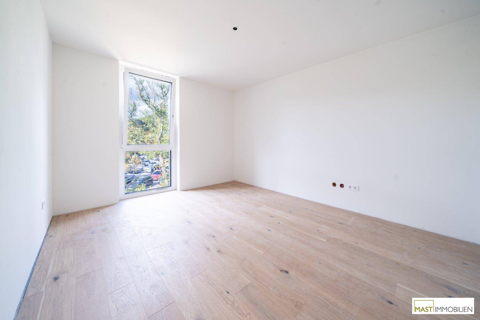 Helles Schlafzimmer mit neuem Holzboden und einem hohen Fenster, das eine grüne Aussicht bietet.