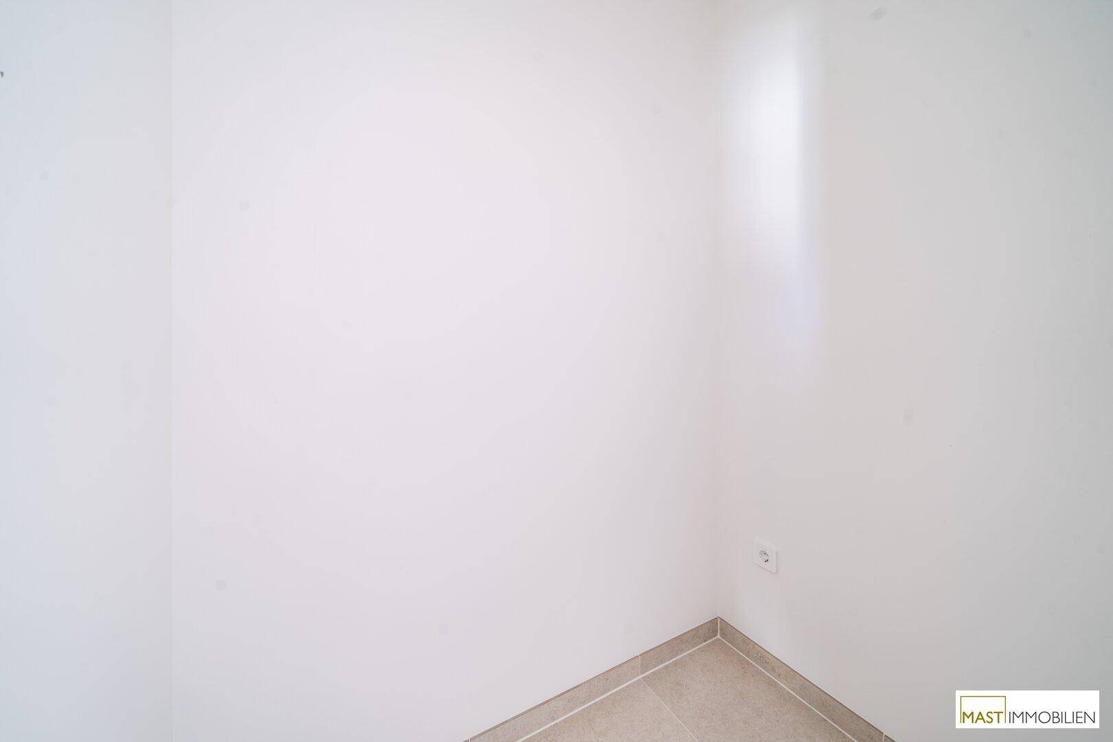 Leere Ecke eines Raumes mit weißen Wänden und hellen Fliesen am Boden.
