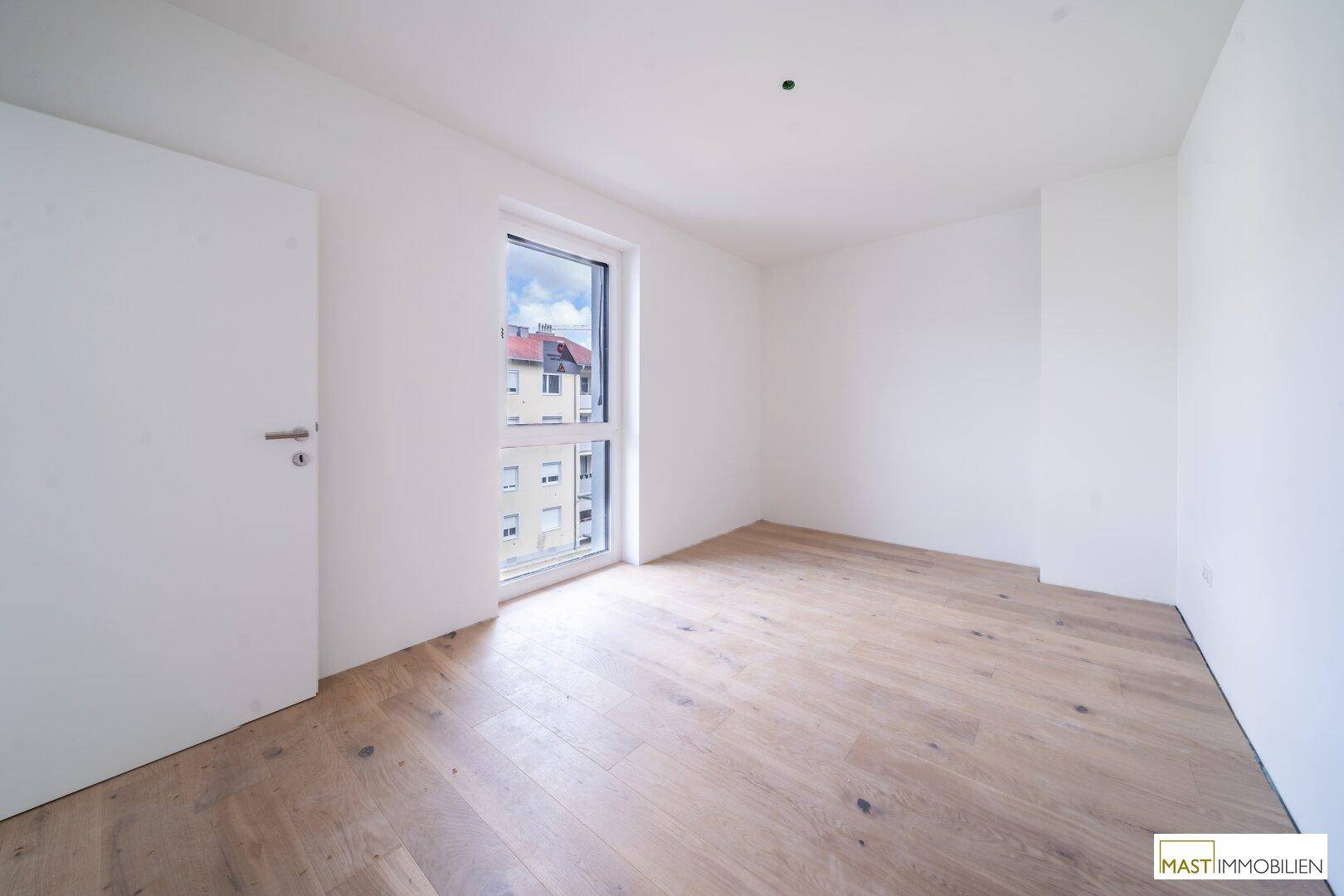 Unmöbliertes Schlafzimmer mit hellem Holzboden und großem Fenster, viel Tageslicht.