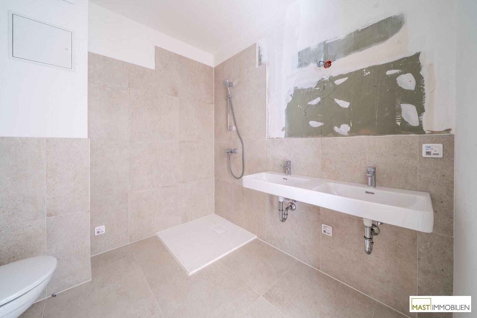 Modernes Badezimmer im Bau mit beigefarbenen Fliesen, Doppelwaschbecken und Duschbereich.