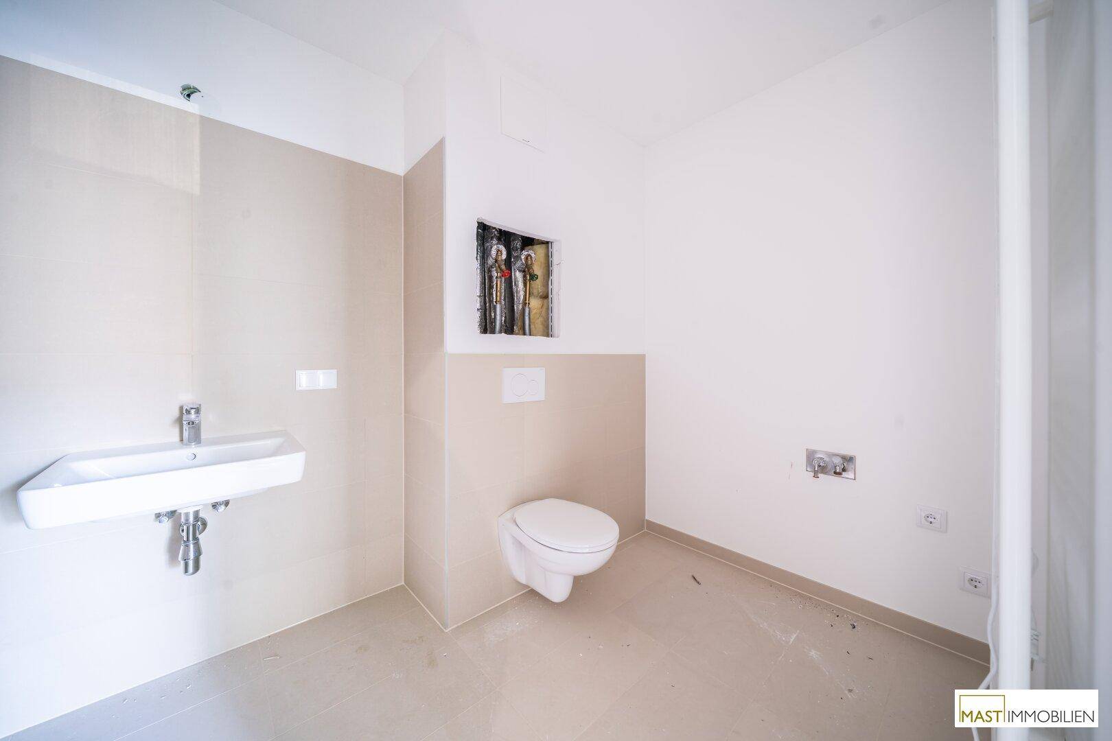 Modernes Badezimmer im Bau mit beigefarbenen Fliesen, Waschbecken und einer wandhängenden Toilette.