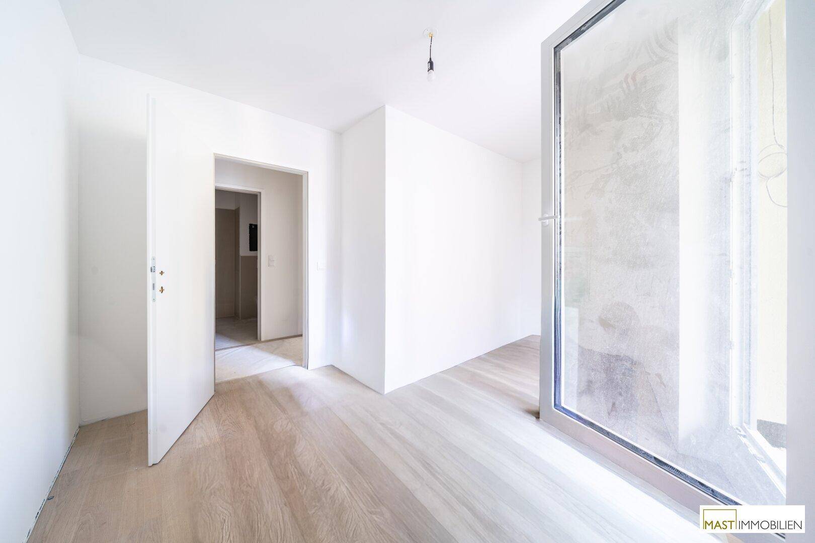 Heller Raum mit neuem Holzboden und großem Fenster, das viel Tageslicht hereinlässt und einen Übergang bietet.