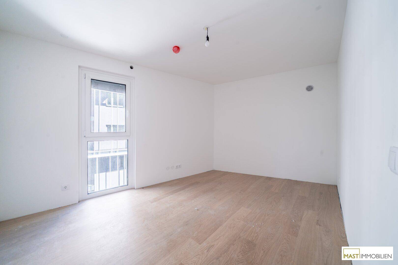 Leeres Schlafzimmer mit hellem Parkettboden und einem großen Fenster, das viel Tageslicht hereinlässt.