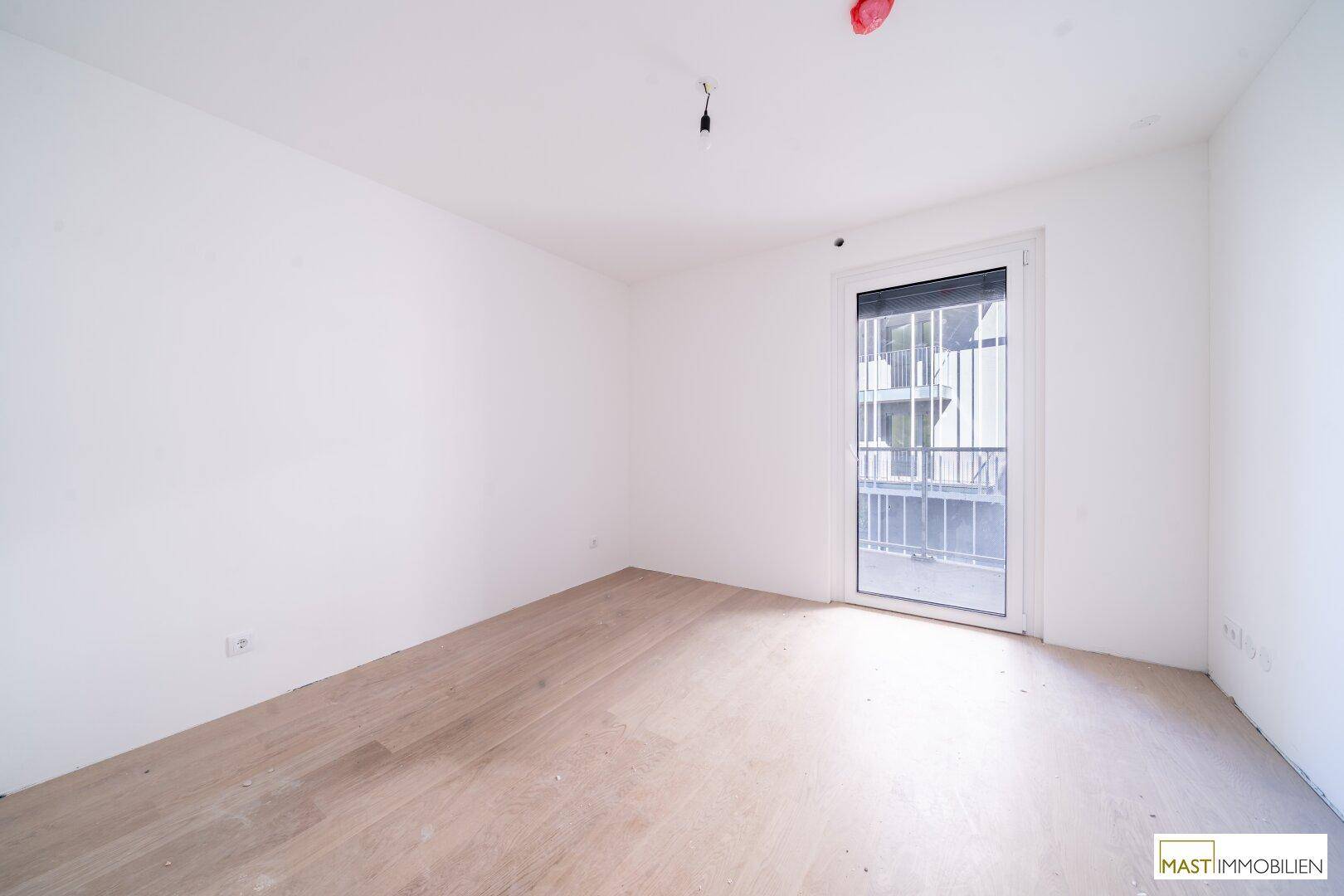 Unmöbliertes Schlafzimmer mit hellem Parkettboden und Zugang zu einem französischen Balkon mit Rollladen.