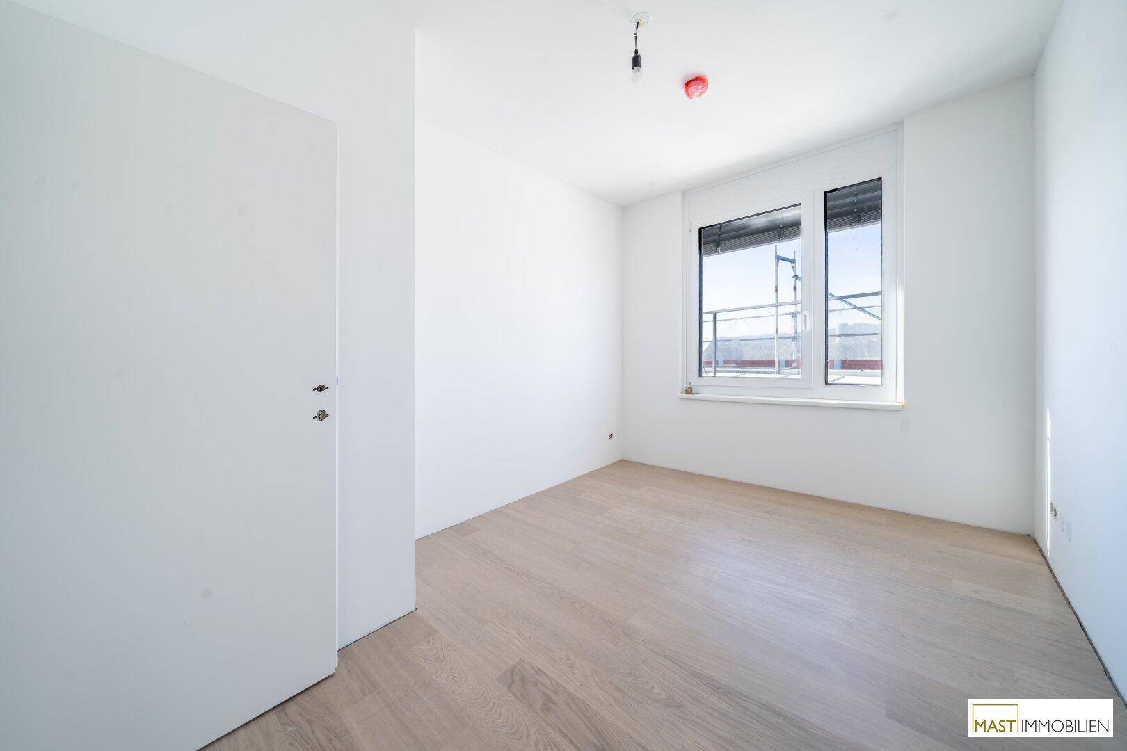 Helles Schlafzimmer mit weiß gestrichenen Wänden, hellem Parkett und einem Fenster mit Ausblick.