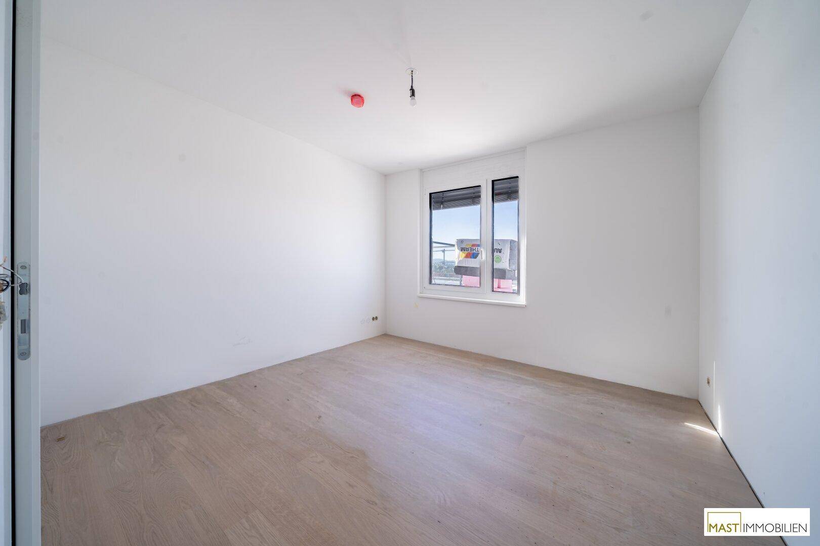Unmöbliertes Schlafzimmer mit großen Fenstern, hellem Holzfußboden und viel Tageslicht.