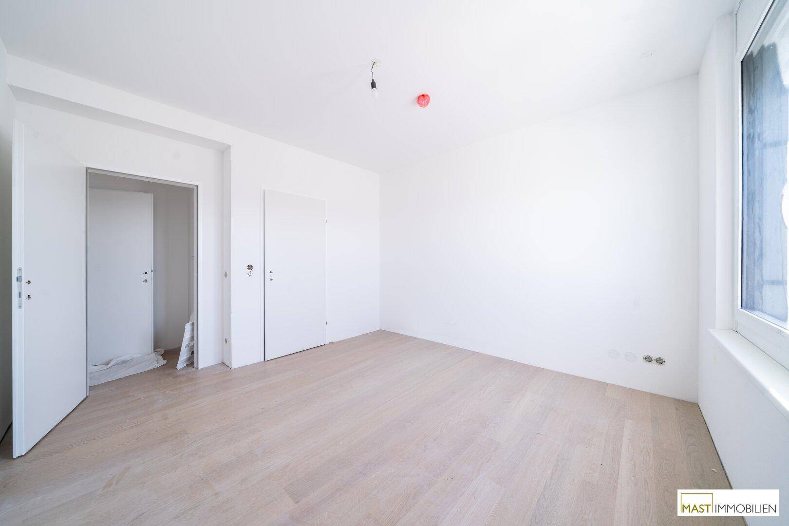 Weiteres helles Zimmer mit weiß gestrichenen Wänden, hellem Parkett und einem Fenster.