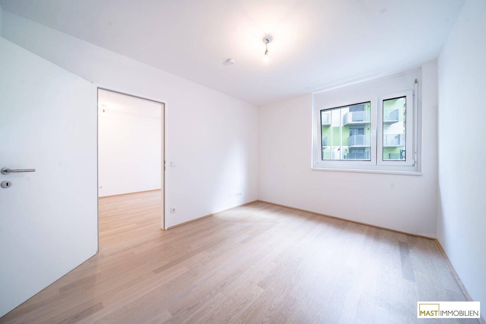 Helles Schlafzimmer mit Laminatboden und einem Fenster, das den Blick auf die grüne Umgebung freigibt.