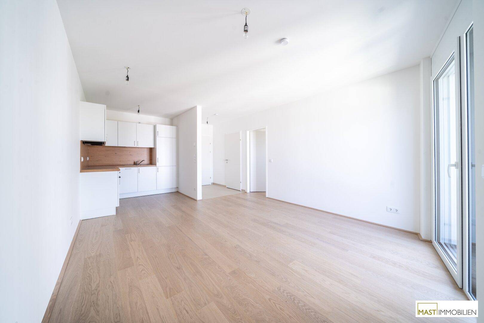 Heller offener Wohnbereich mit integrierter Küchenzeile, Parkettboden und Zugang zum Balkon.
