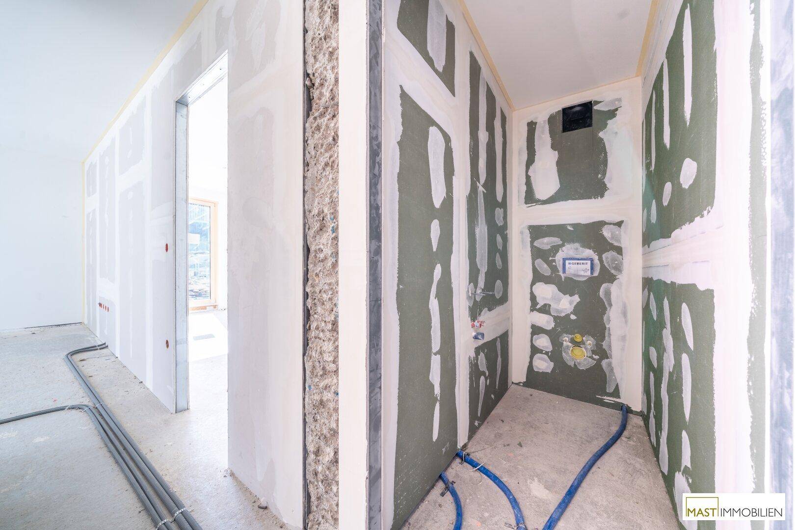 Flur im Rohbau, der zu einem Badezimmer führt, mit sichtbaren Installationen und Trockenbauwänden.