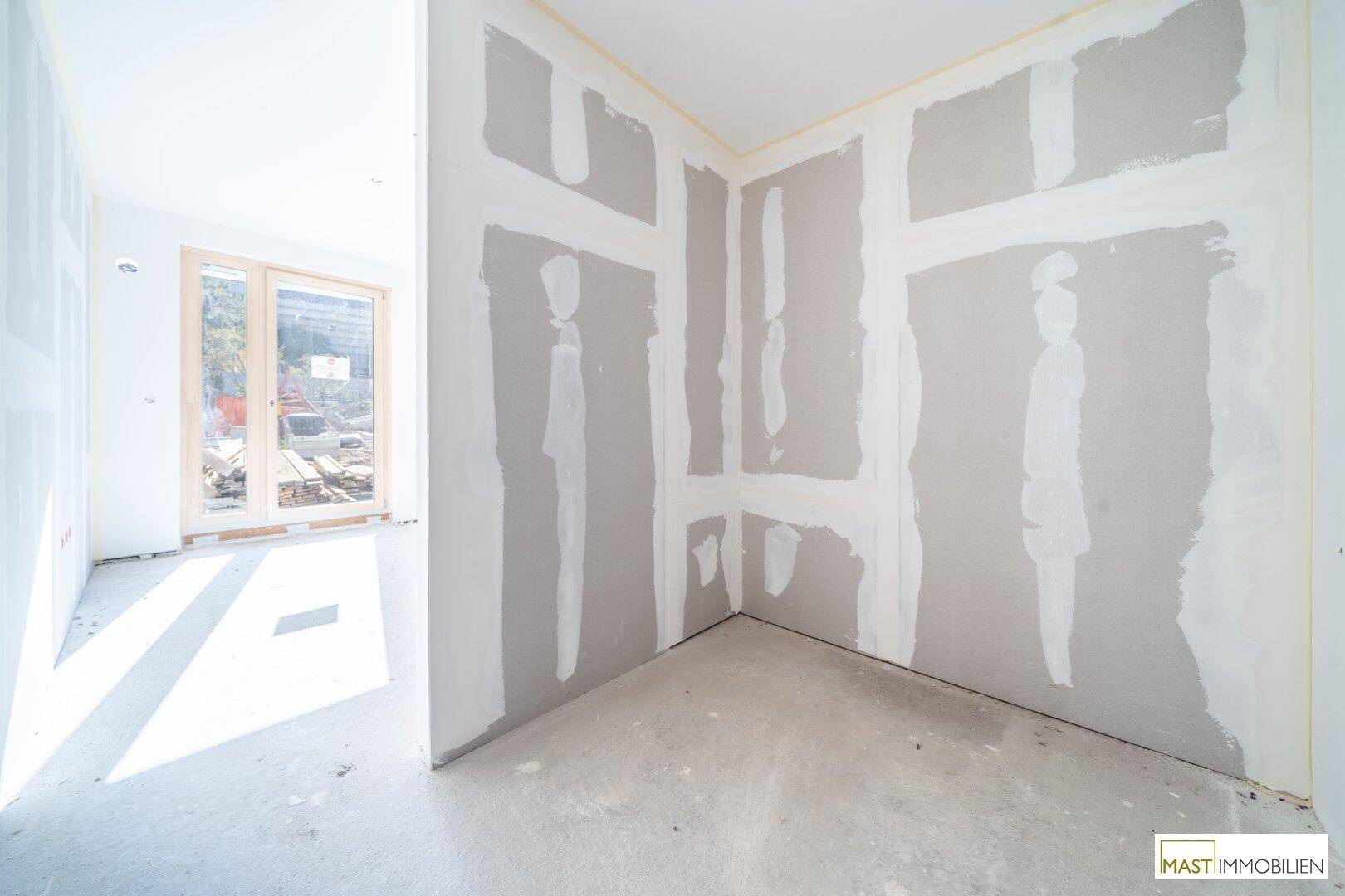 Ecke eines Rohbauraumes mit vorbereiteten Wänden und viel Tageslicht durch ein angrenzendes Fenster.
