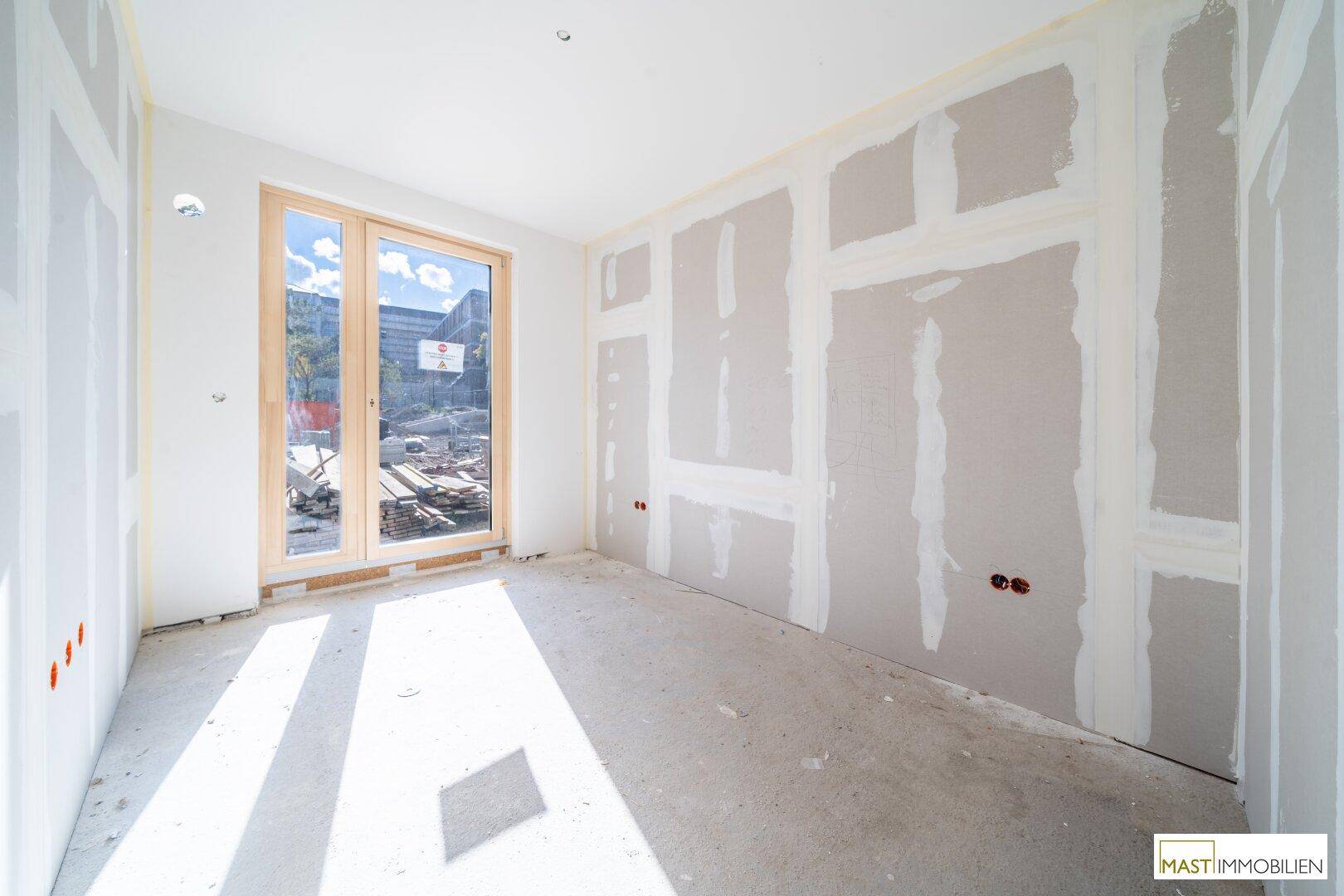 Heller Rohbauraum mit einem großen Fenster, das viel Sonnenlicht hereinlässt und den Baufortschritt zeigt.