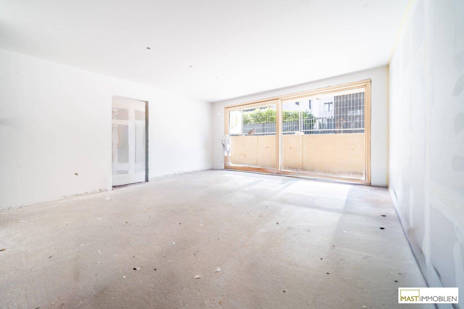 Unfertiger Raum mit Rohbauwänden, Betonboden und Zugang zu einem Balkon oder einer Terrasse.
