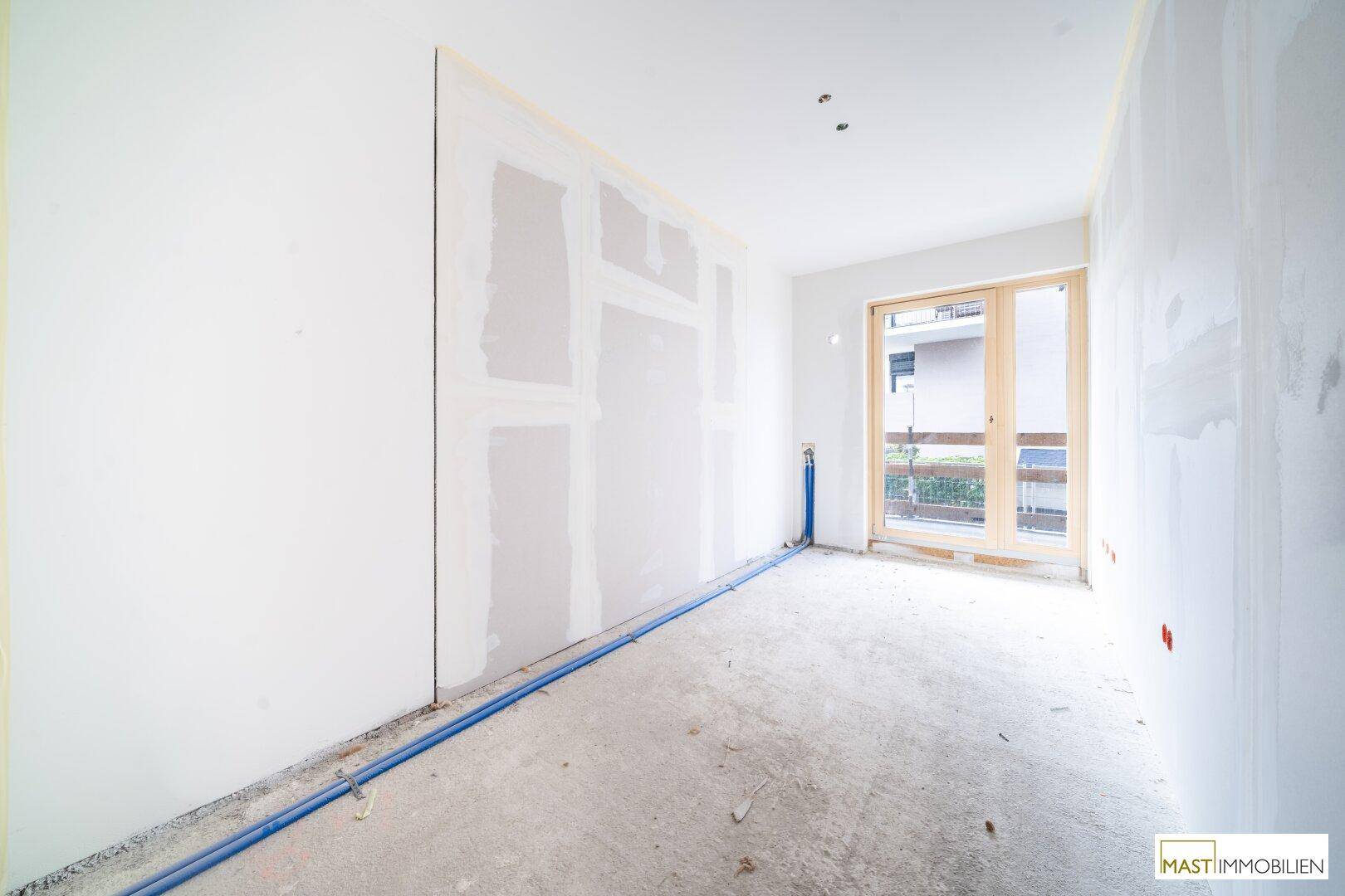 Unfertiges Schlafzimmer mit sichtbaren Rohrleitungen, Trockenbauwänden und großem Fenster.