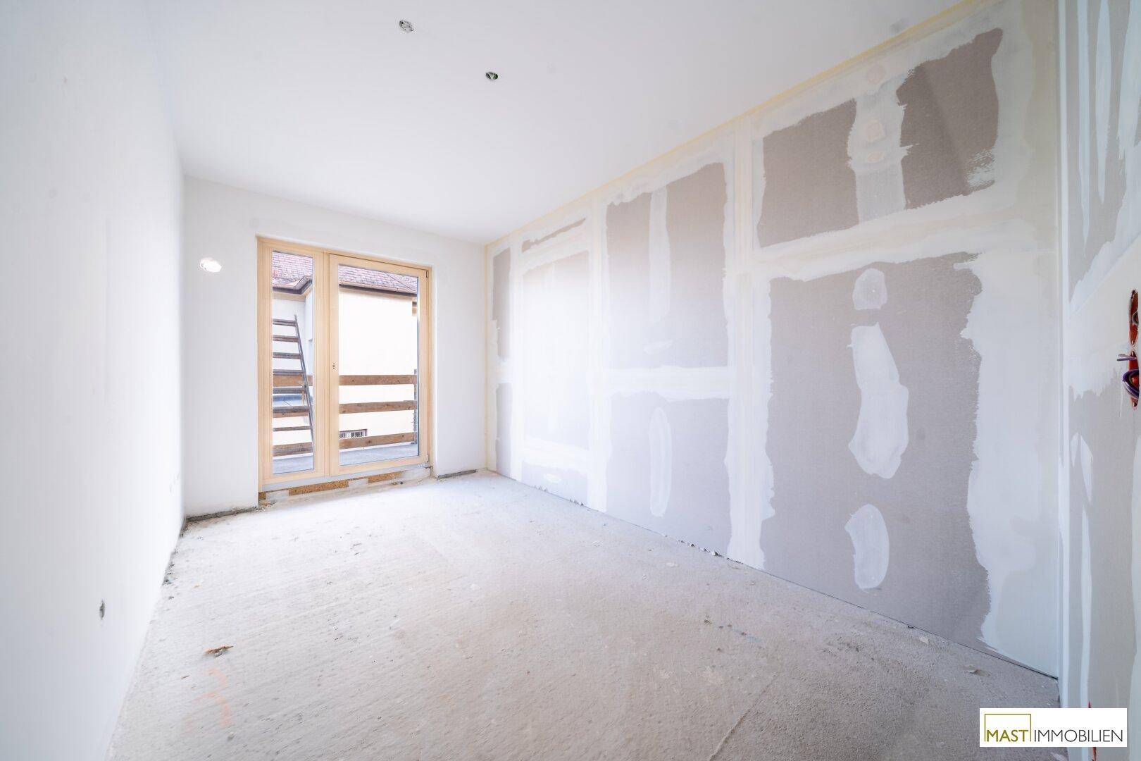 Unfertiges Zimmer mit Trockenbauwänden und Balkonzugang, bereit für den Ausbau.