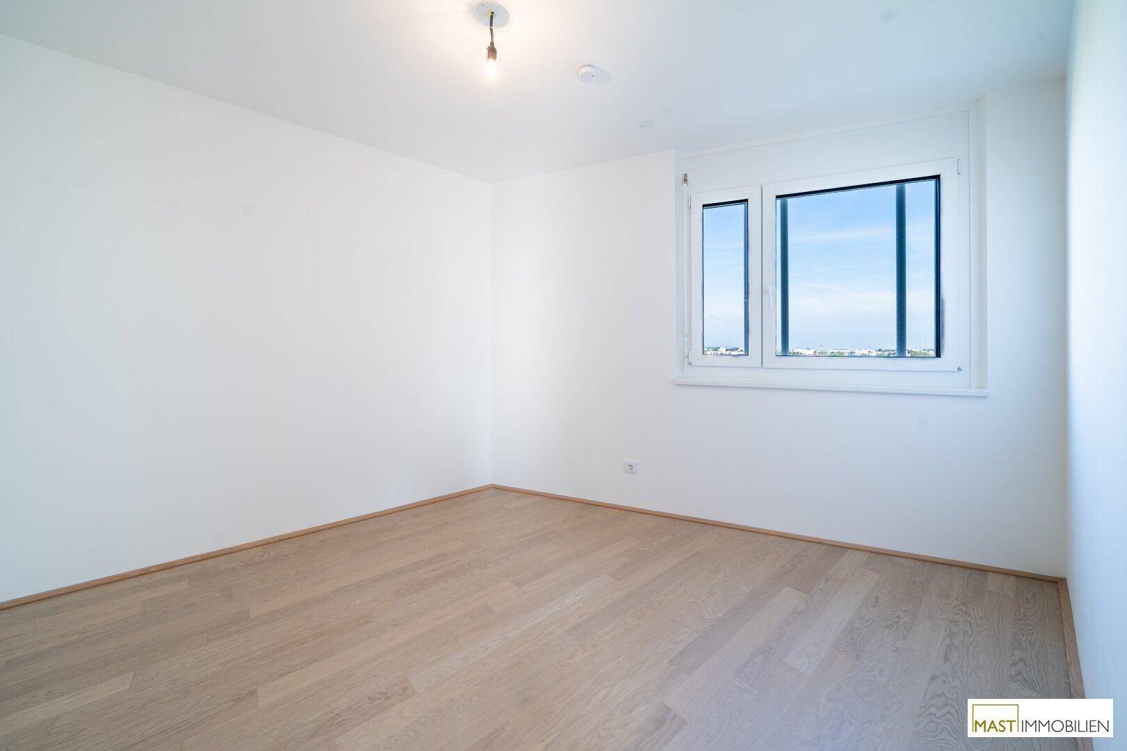Leeres Schlafzimmer mit hellem Parkettboden und großem Fenster, bietet viel natürliches Licht.