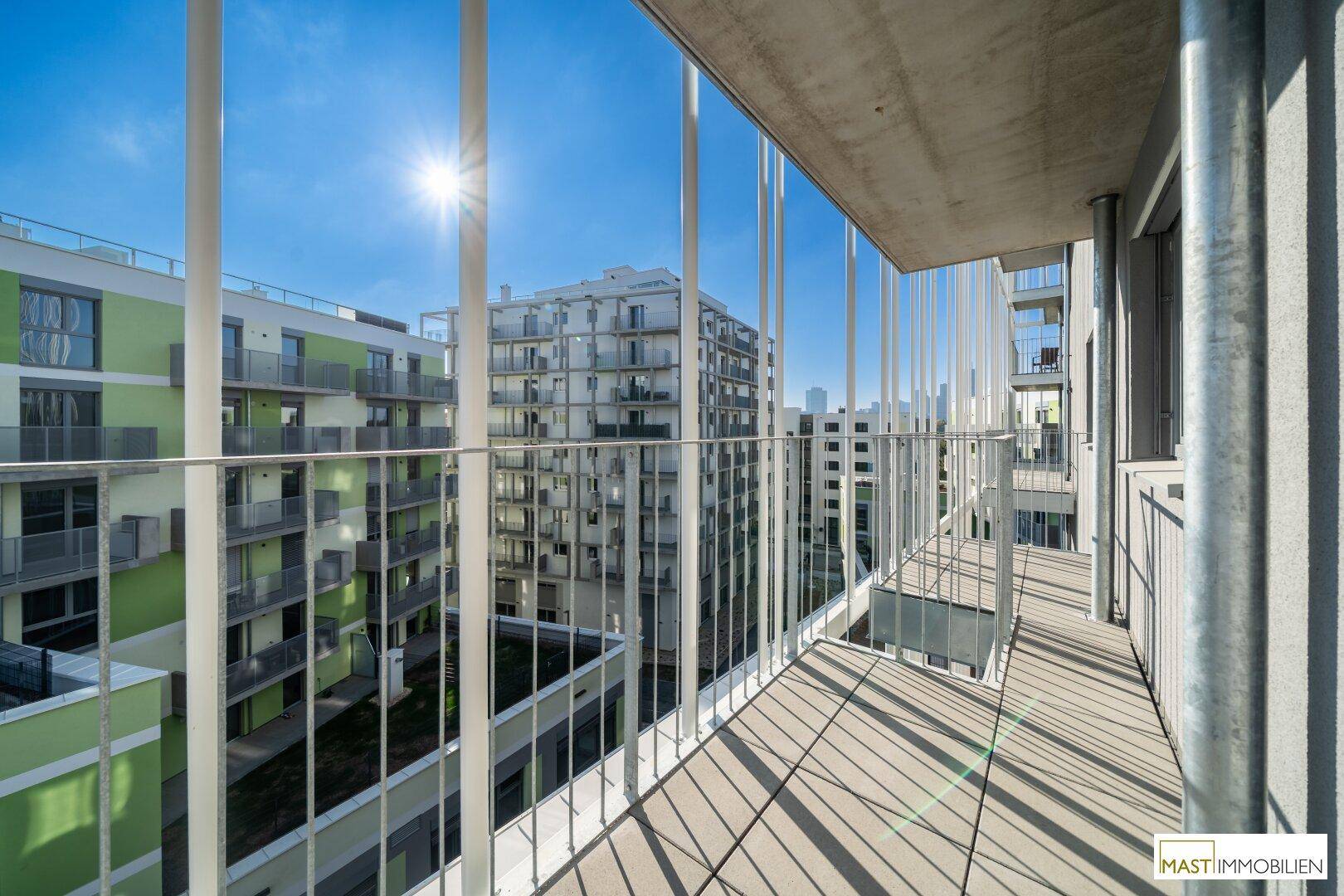 Langer Balkon mit Fliesenboden und Blick auf moderne Wohngebäude, sonnig und offen.
