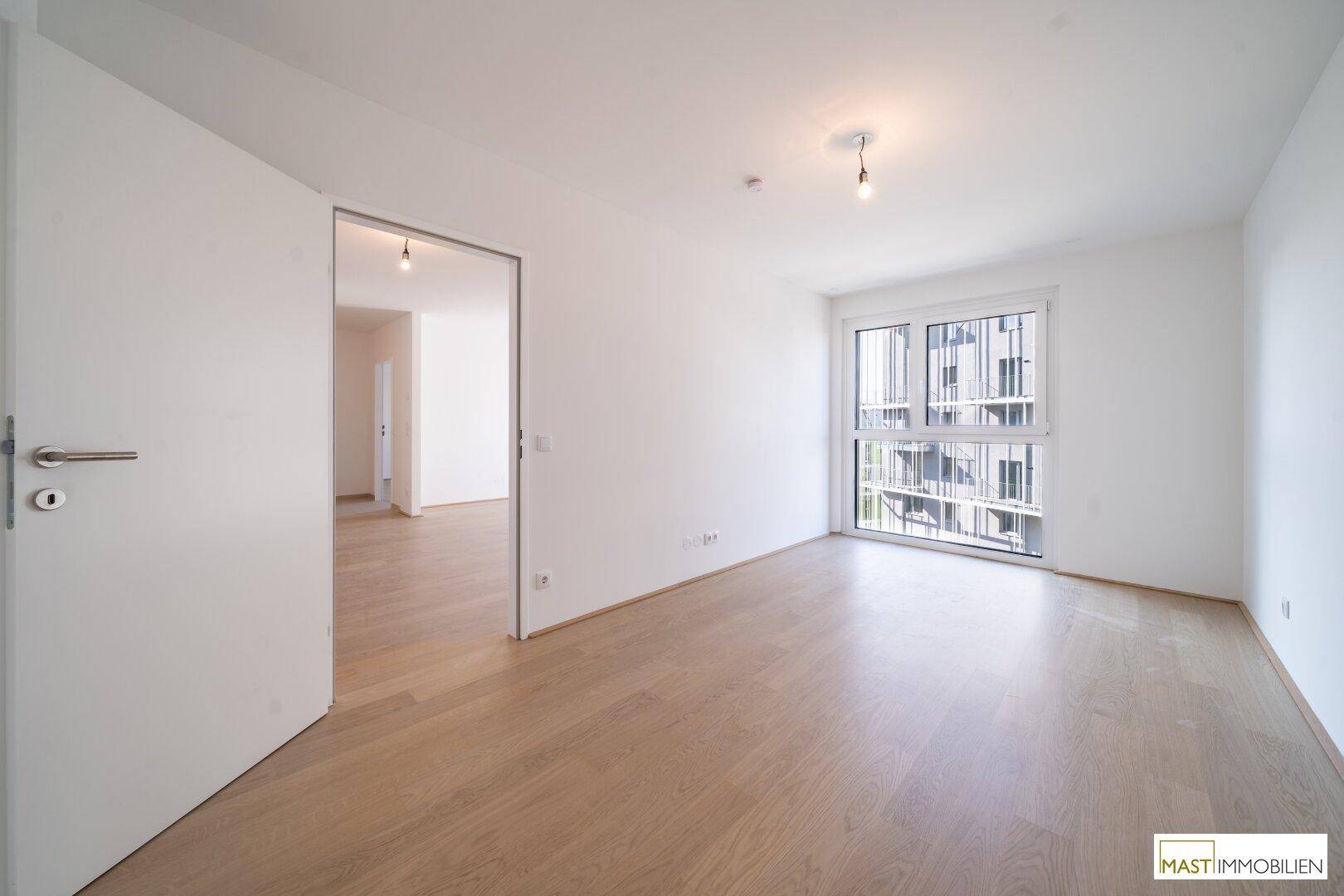 Leeres Schlafzimmer mit hellem Parkettboden und einem großen Fenster mit Blick nach draußen.