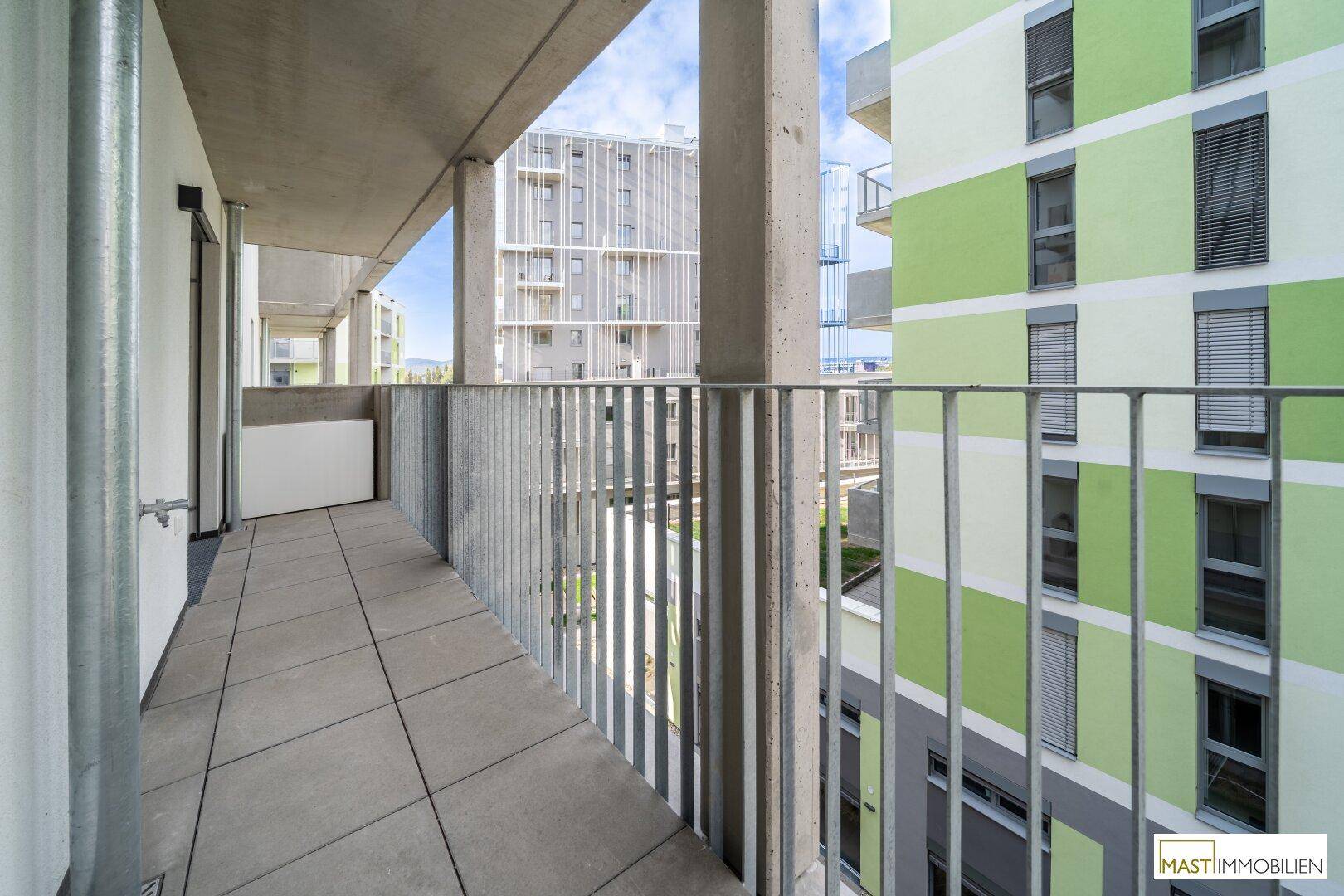 Balkon mit Metallgeländer und Blick auf die grüne Fläche und benachbarte Gebäude der Wohnanlage.