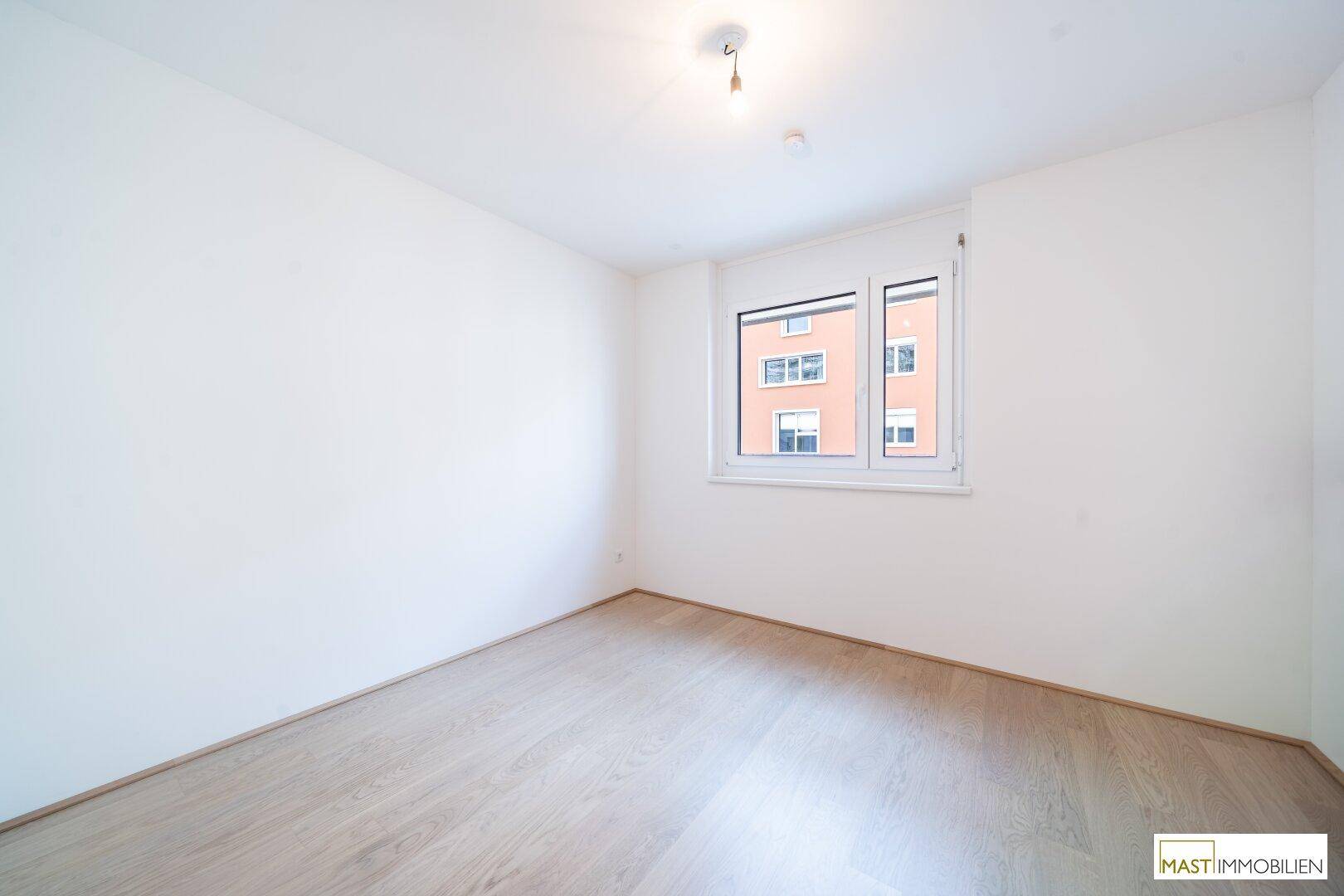 Helles, leeres Zimmer mit hellem Laminatboden und einem Fenster mit Blick auf ein Gebäude.