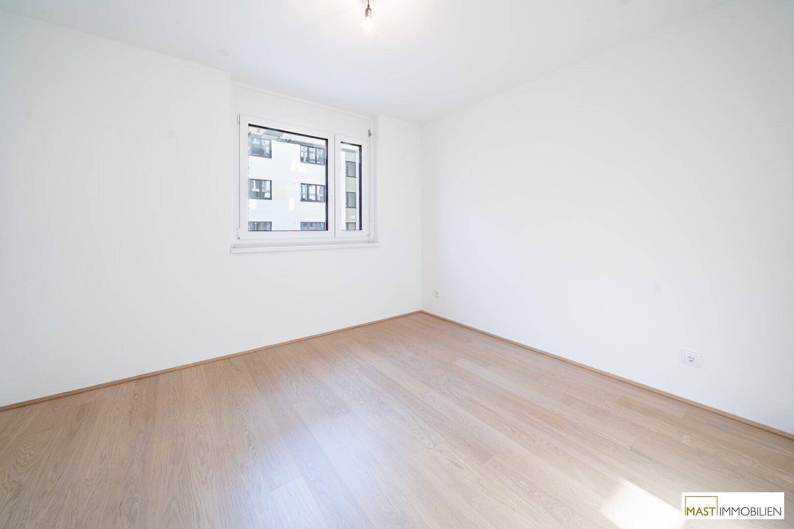 Leeres Zimmer mit hellem Laminatboden und einem Fenster, das Tageslicht hereinlässt.