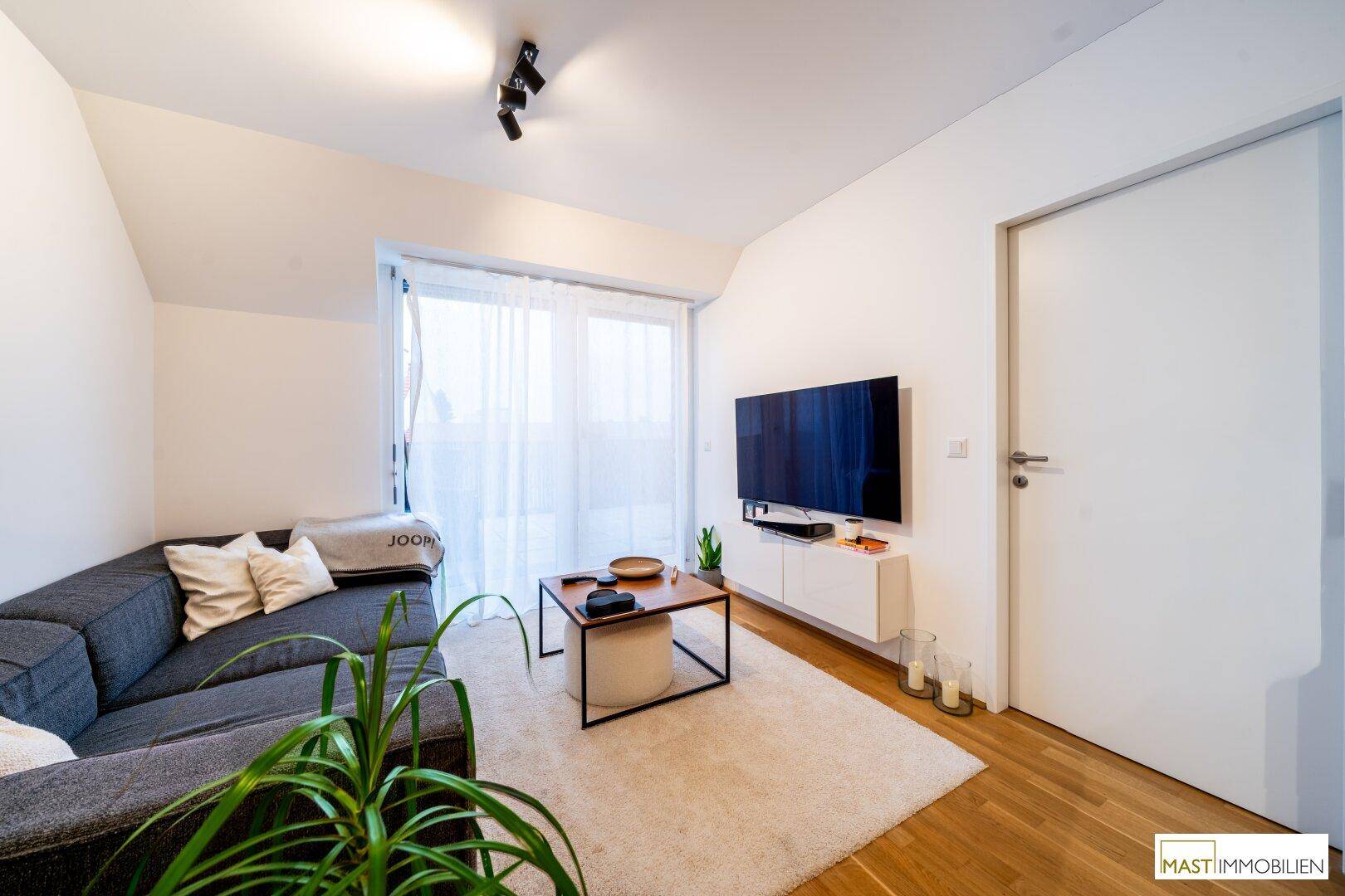 Gemütlicher Wohnbereich mit grauem Sofa, Fernseher und hellem Teppichboden.