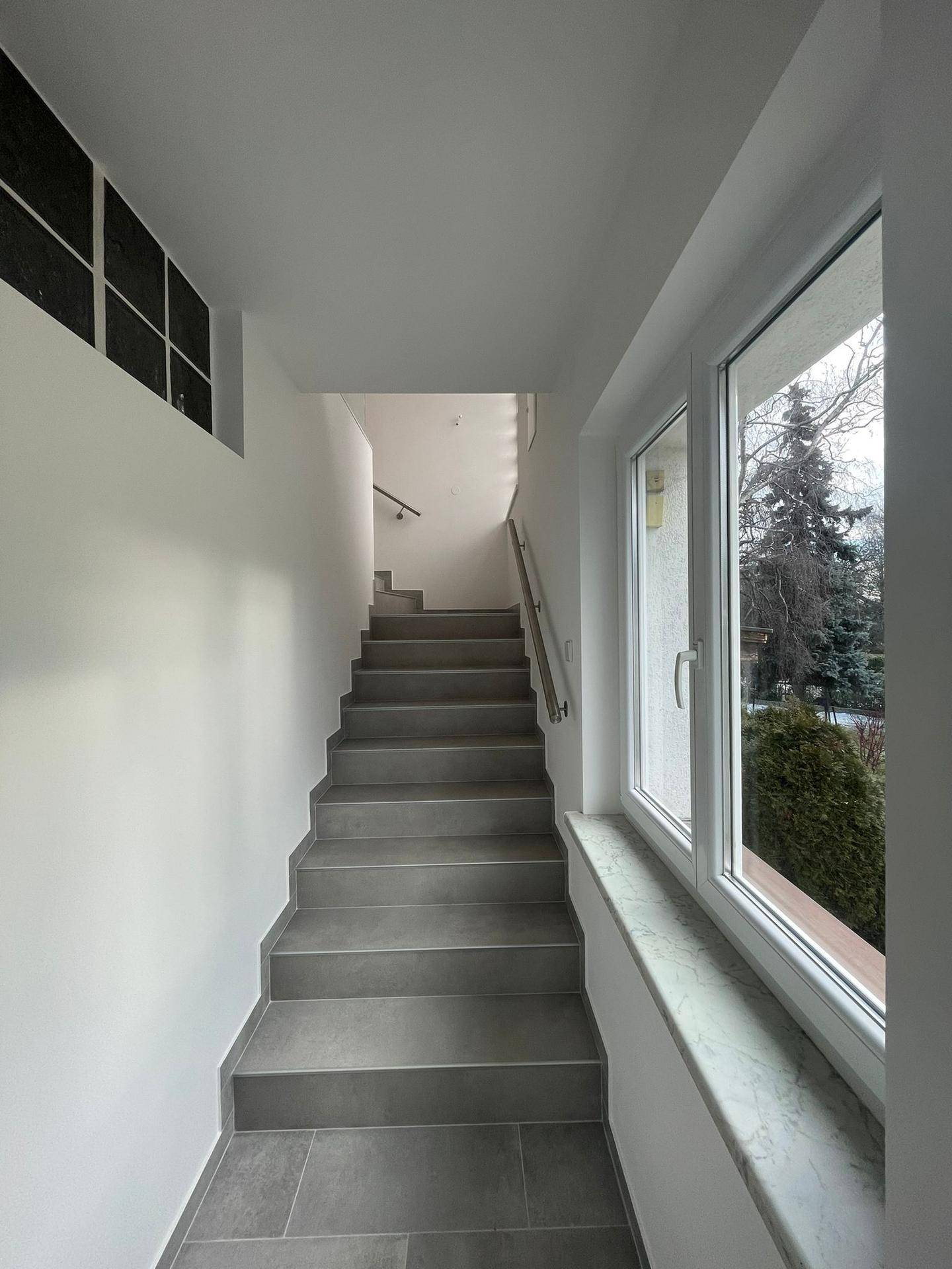 Treppenhaus mit grauen Fliesen und einem Fenster, das den Bereich mit Tageslicht erhellt.