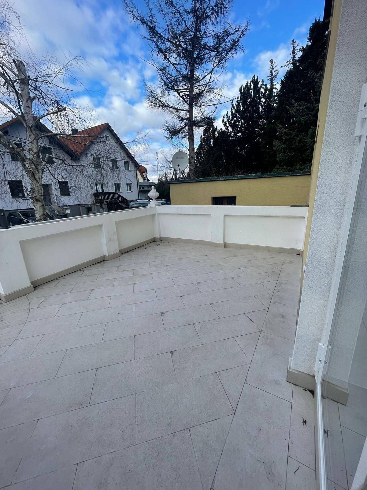 Geräumige Außenterrasse mit hellen Fliesen und einer niedrigen Mauer, umgeben von Bäumen.