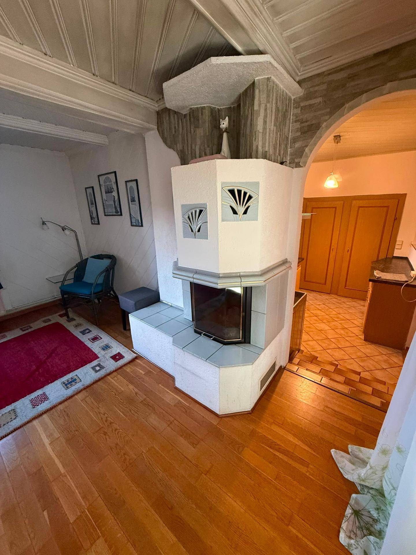 Wohnzimmer mit Kachelofen, Holzboden und Zugang zu einem weiteren Raum.