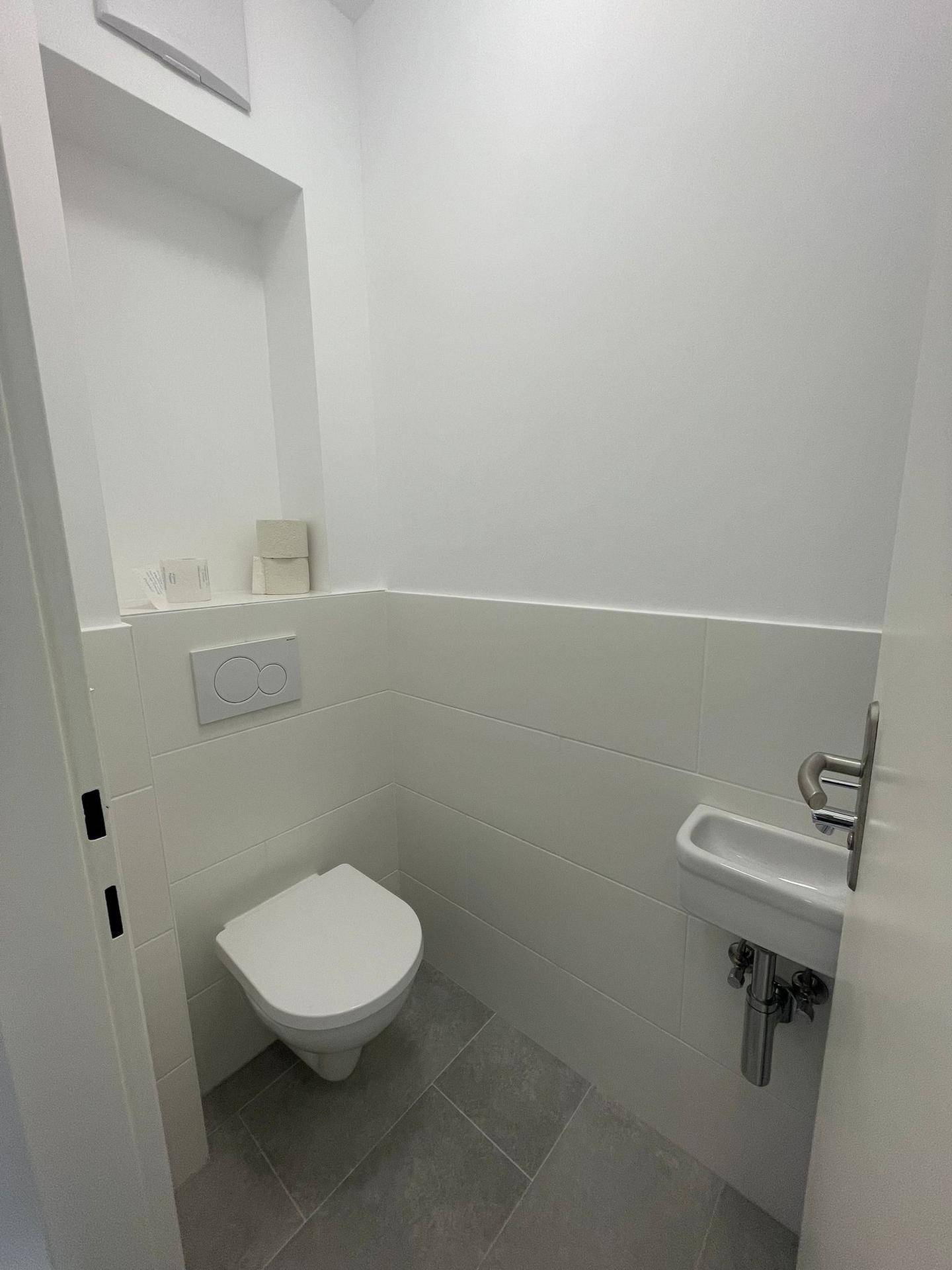 Modernes Gäste-WC mit wandhängender Toilette, kleinem Waschbecken und hellen Fliesen.