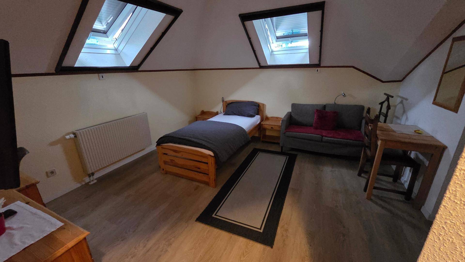 Helles Dachgeschosszimmer mit Einzelbett, Sofa, kleinem Tisch und zwei Dachfenstern.
