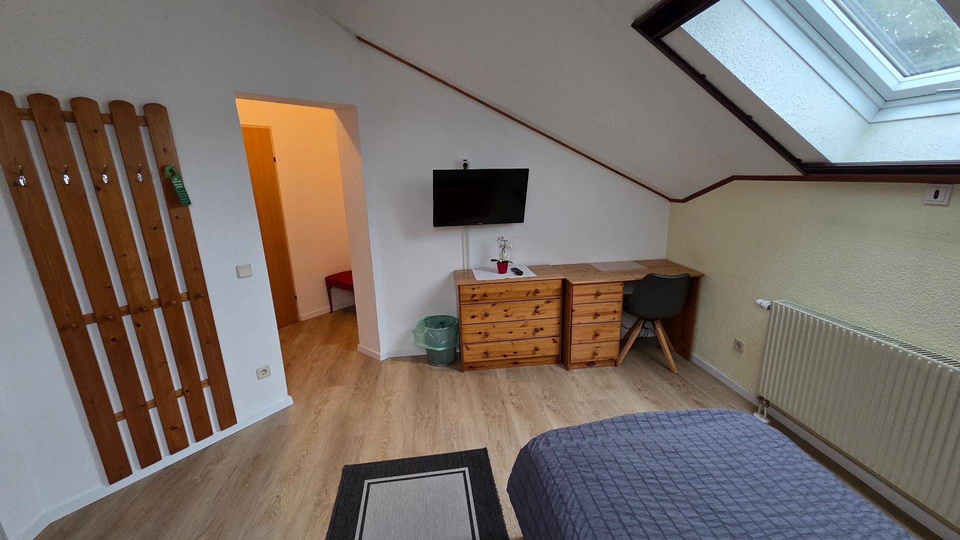 Dachgeschosszimmer mit Schreibtisch, Wandfernseher, Garderobe und Zugang zu einem weiteren Raum.