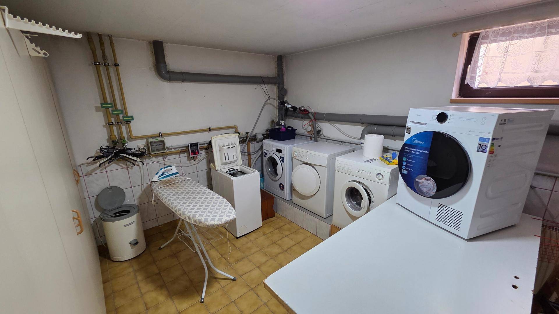 Hauswirtschaftsraum mit Waschmaschinen, Trockner, Bügelbrett und sichtbaren Leitungen.