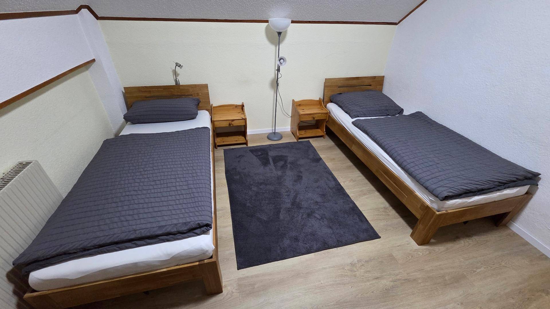 Zwei Einzelbetten aus Holz mit grauer Bettwäsche, Nachttischen und einer Stehlampe.