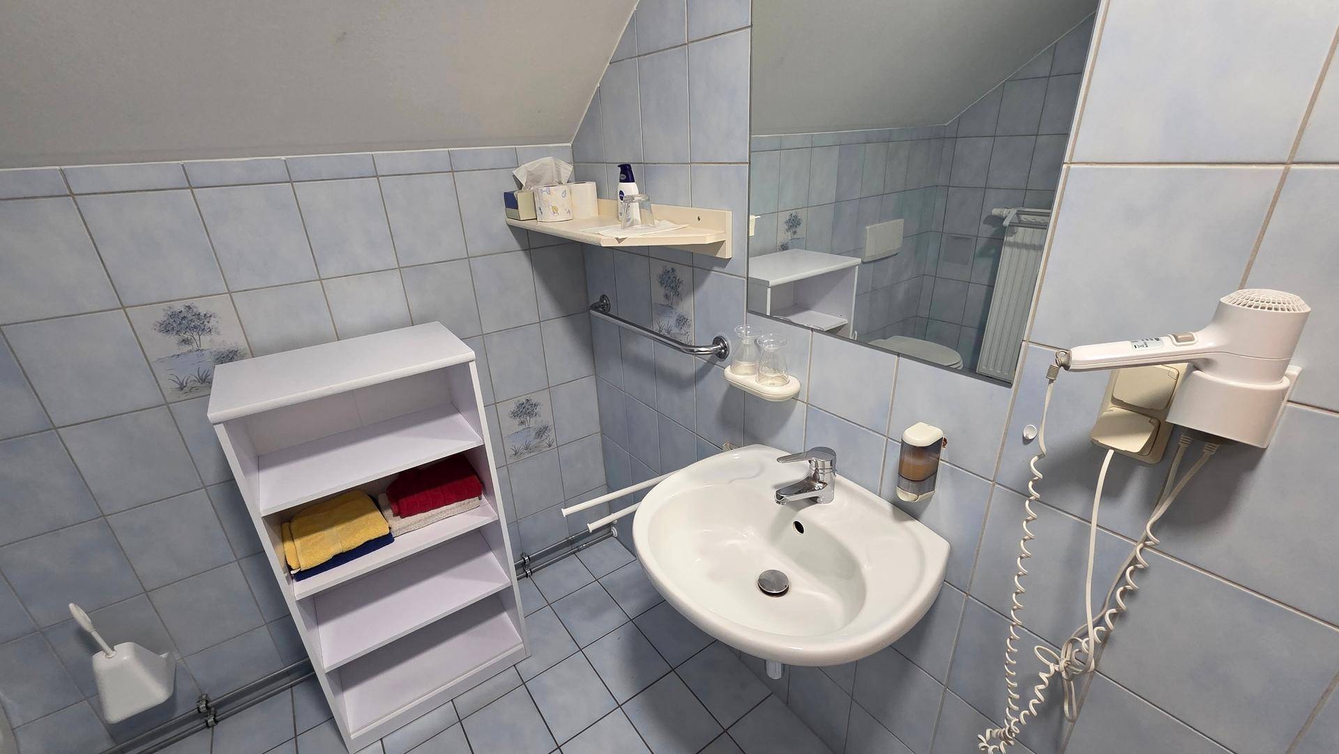 Badezimmer mit blauen Fliesen, Waschbecken, Spiegel, Föhn und kleiner Ablage.