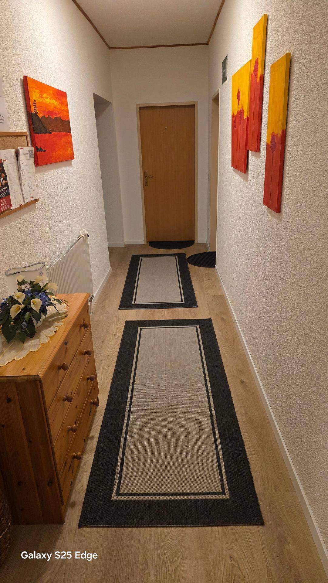 Heller Flur mit Holzfußboden, Kommode und farbenfrohen abstrakten Bildern an der Wand.