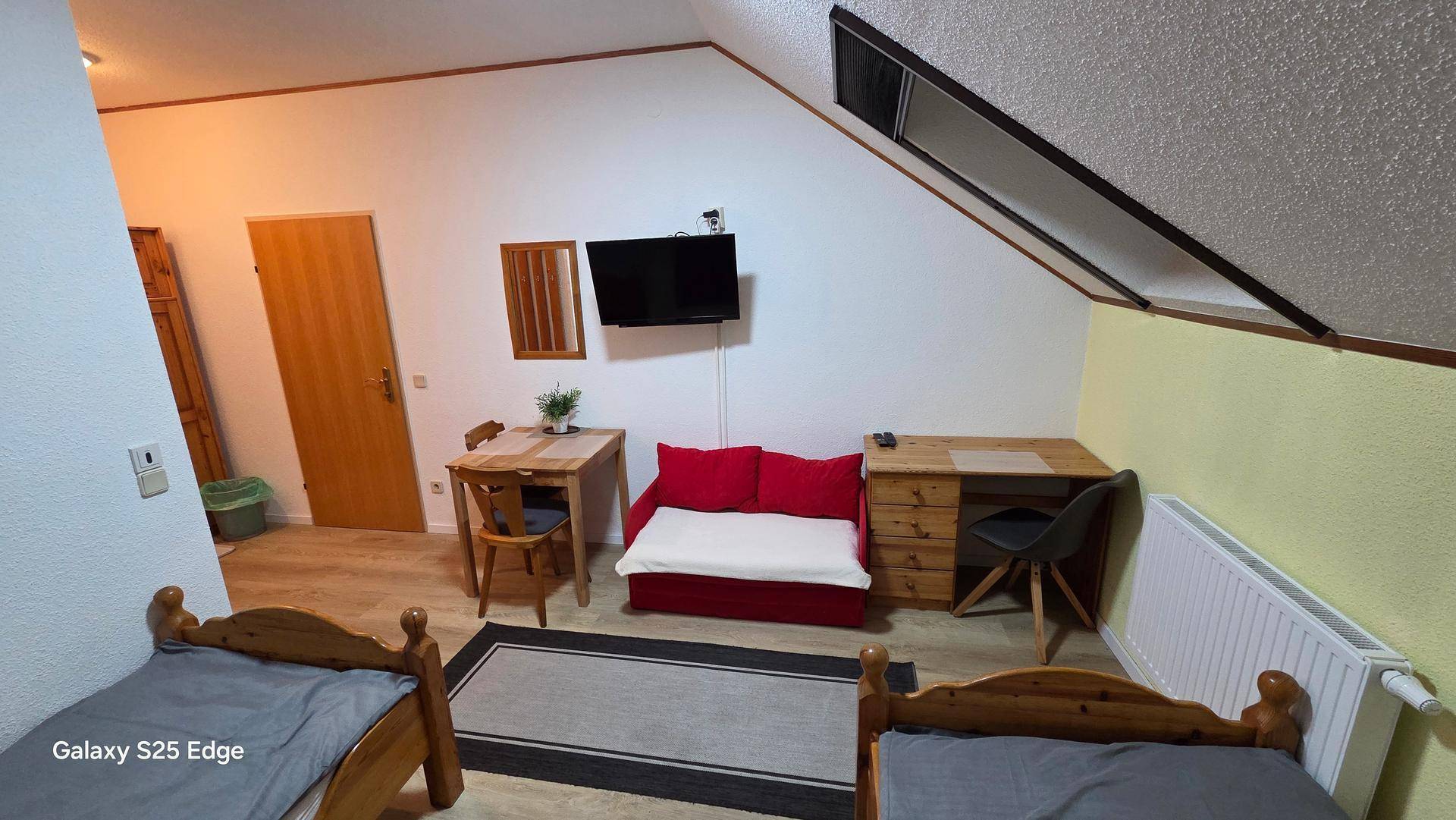 Dachgeschosszimmer mit zwei Einzelbetten, rotem Schlafsofa, Esstisch und Wandfernseher.