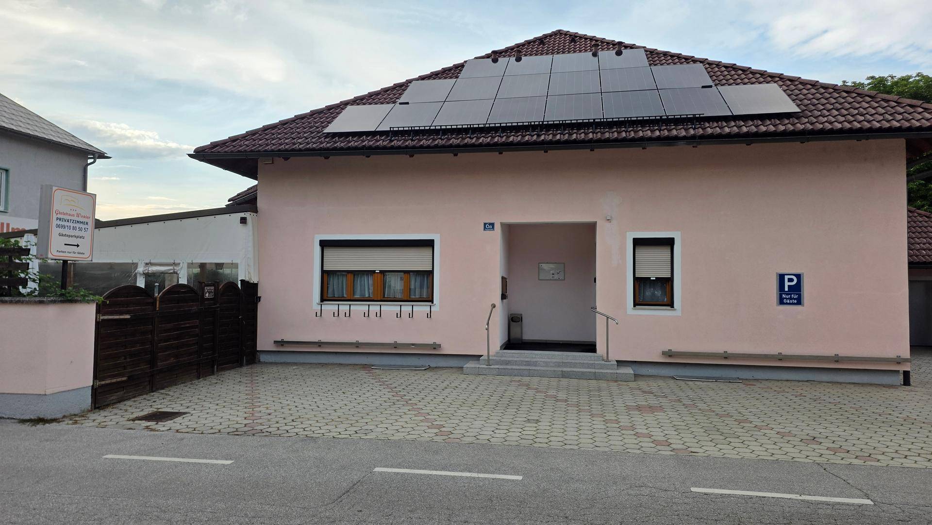 Rosa Gebäude mit Ziegeldach, Solarpaneelen und einem kleinen Eingangsbereich mit Parkplatz.