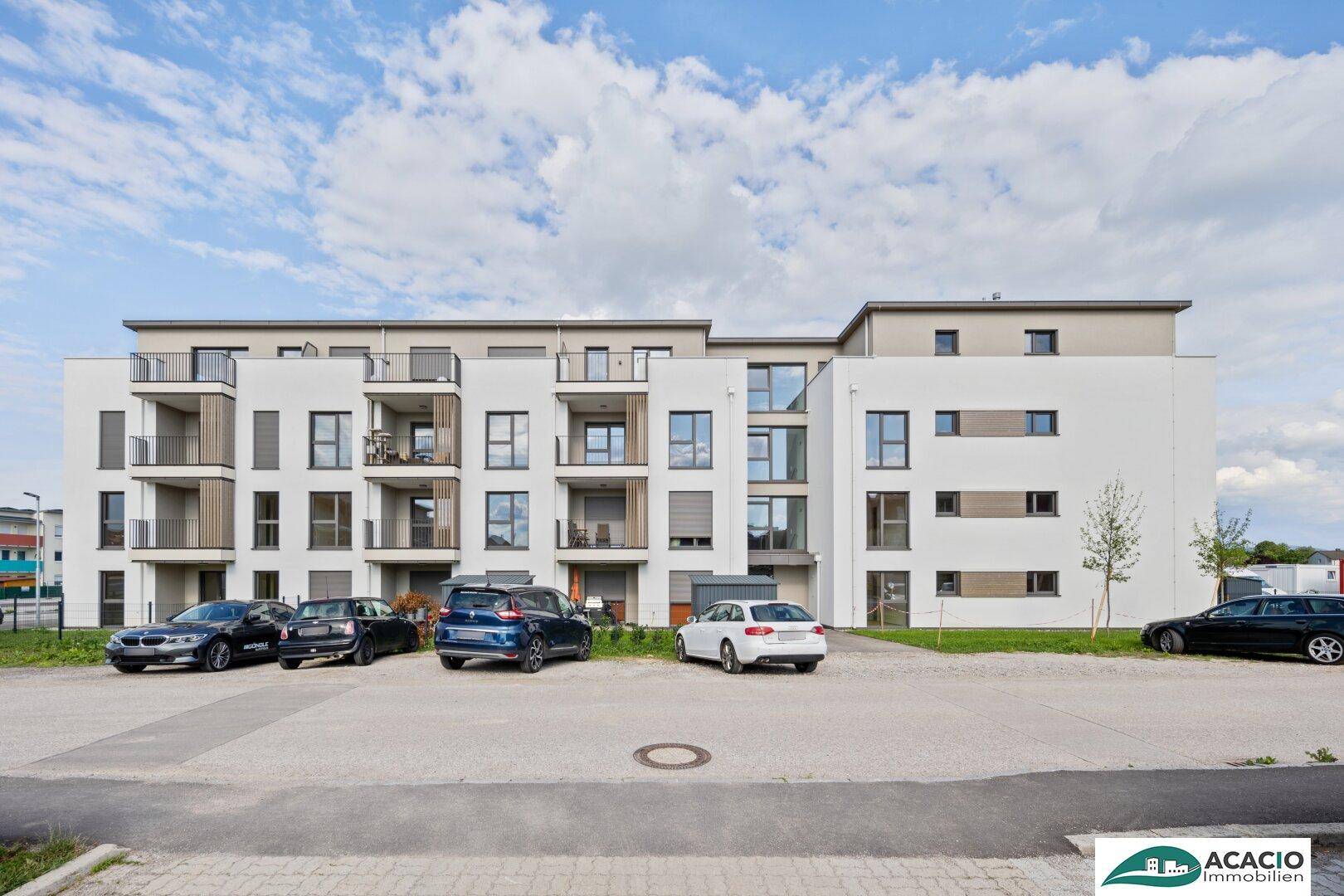 Modernes Mehrfamilienhaus mit weißer Fassade, mehreren Balkonen und Außenstellplätzen unter blauem Himmel.