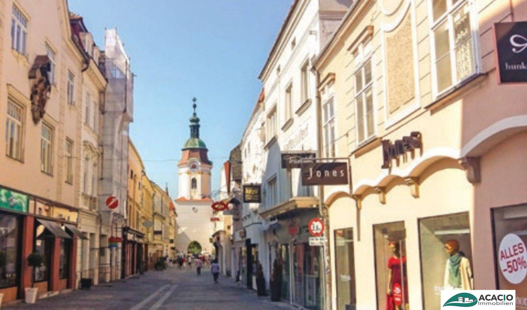 Belebte Einkaufsstraße mit historischen Gebäuden und einem Kirchturm im Hintergrund unter blauem Himmel.
