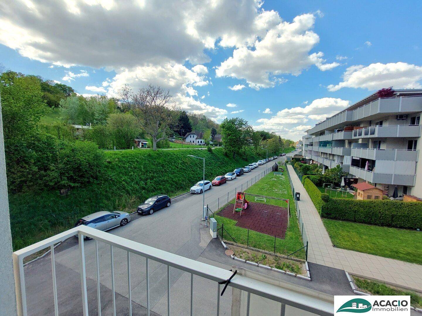 Balkon mit Blick auf eine ruhige Straße, einen Spielplatz und die umliegende grüne Landschaft.