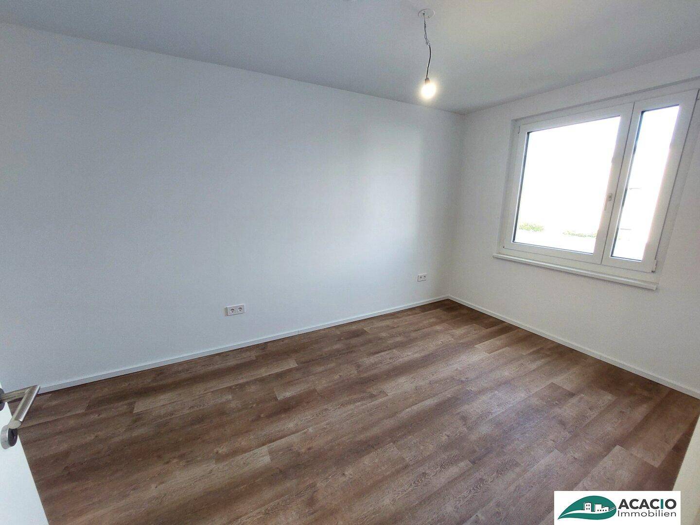 Leeres Schlafzimmer mit Laminatboden, weißen Wänden und viel Tageslicht durch das Fenster.